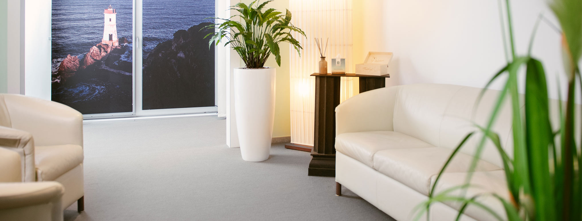 Heller Empfangsbereich mit weißen Sofas, einer großen Pflanze und einem Wandbild mit Leuchtturm-Motiv.