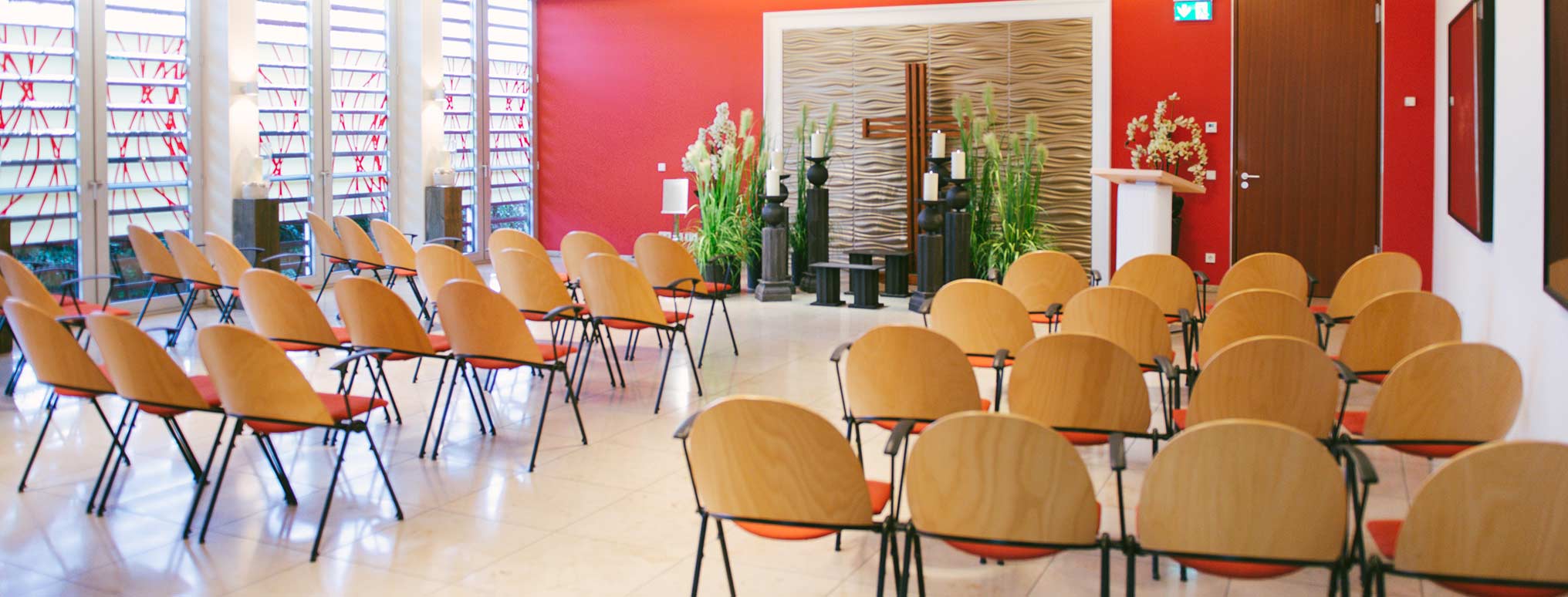 Moderne Trauerhalle mit roten Wänden, kunstvoll gestalteten Fenstern und Reihen von Stühlen für die Trauerfeier.